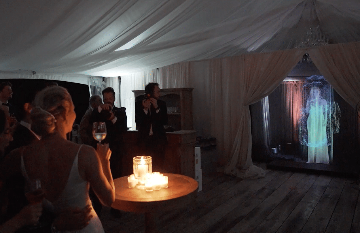 Photo shows a bridesmaid giving a toast as a hologram at a wedding.
